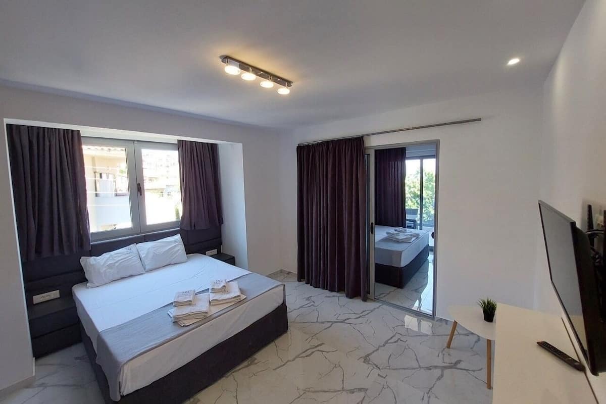 A modern hotel room with a large bed, a desk with a computer, and a balcony with a view.