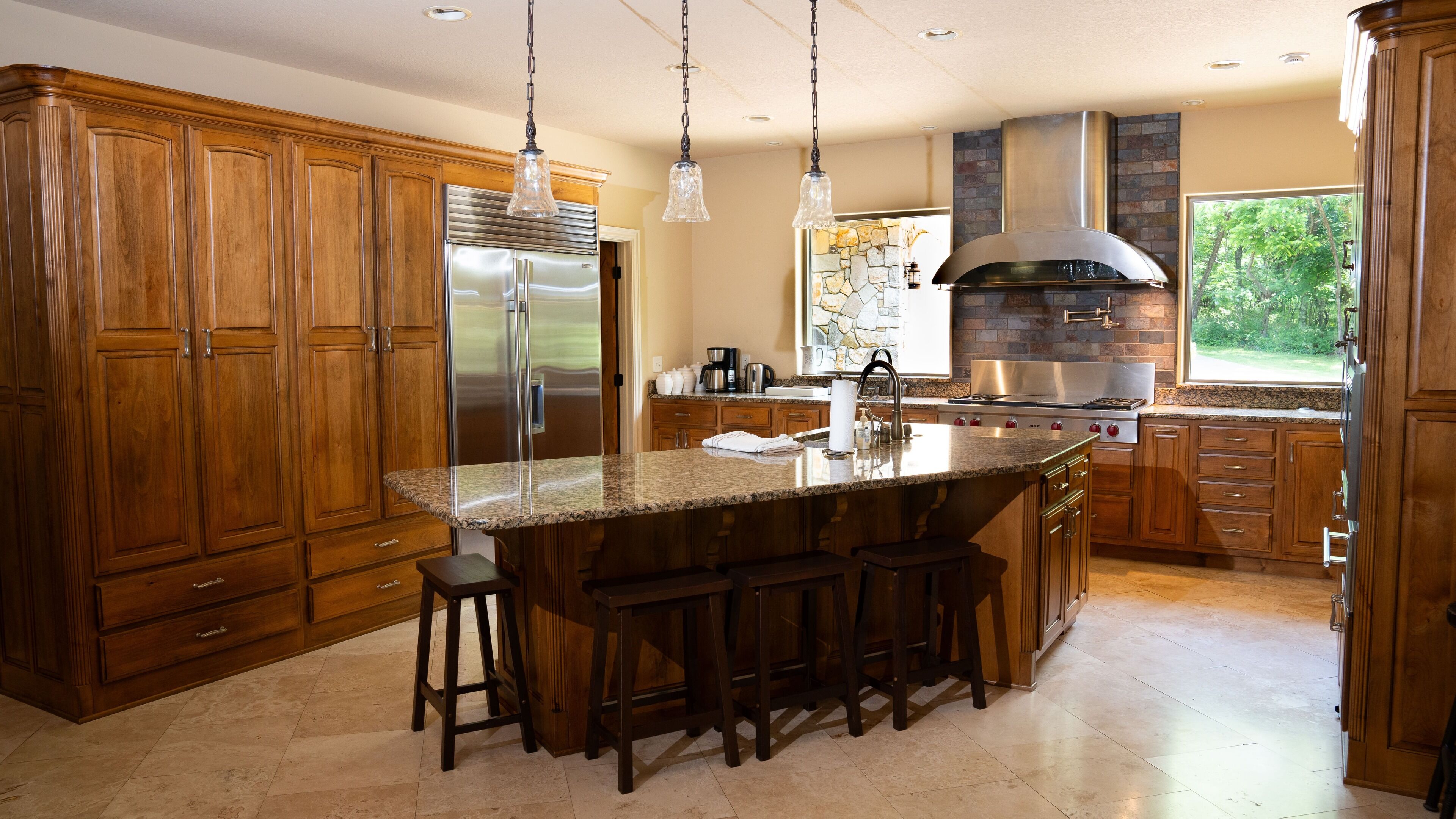 A spacious kitchen with wooden cabinets, a large island with bar stools, stainless steel appliances, and a stone backsplash.