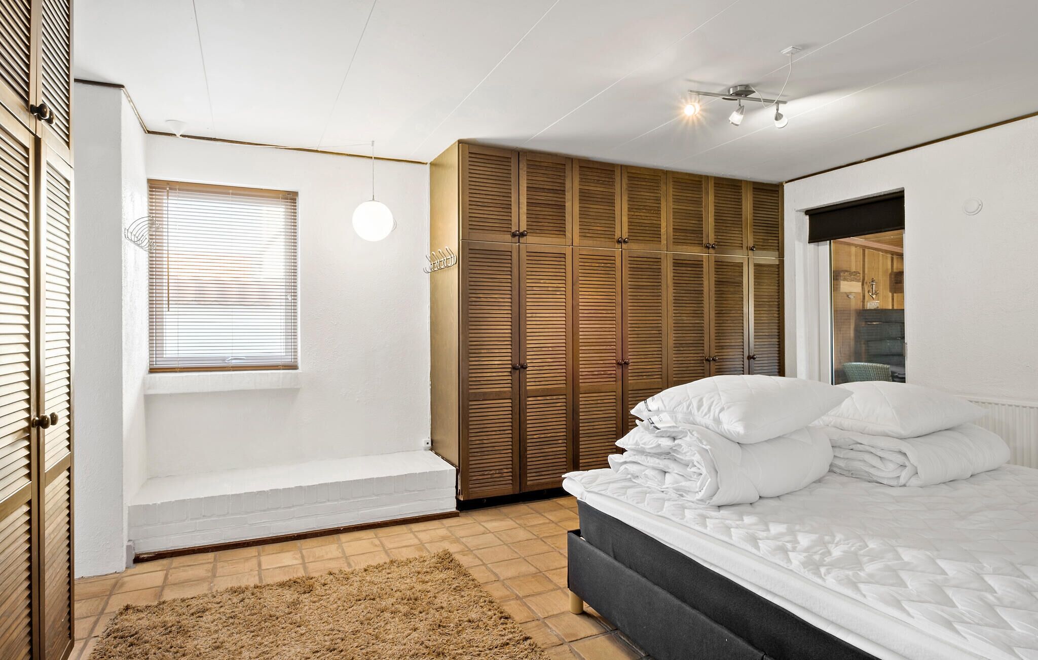 A bedroom with a bed, a window with blinds, a wooden wardrobe, and a bench.