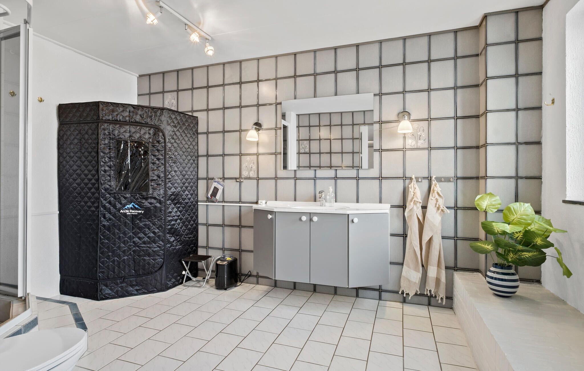 Une salle de bain moderne avec une cabine de douche noire à motifs matelassés, des meubles gris, un lavabo blanc et un miroir au-dessus. Deux serviettes sont accrochées à des crochets et une plante en pot se trouve sur la droite.