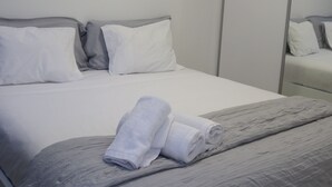 Una cama bien hecha con ropa de cama blanca y gris, una toalla enrollada y una manta gris con textura.