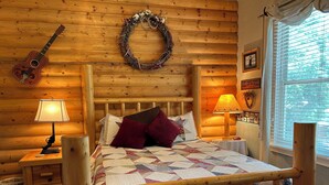 Une chambre chaleureuse dans un chalet en rondins, avec un lit en bois, deux lampes de chevet, une guitare, une couronne décorative et une fenêtre avec des stores.
