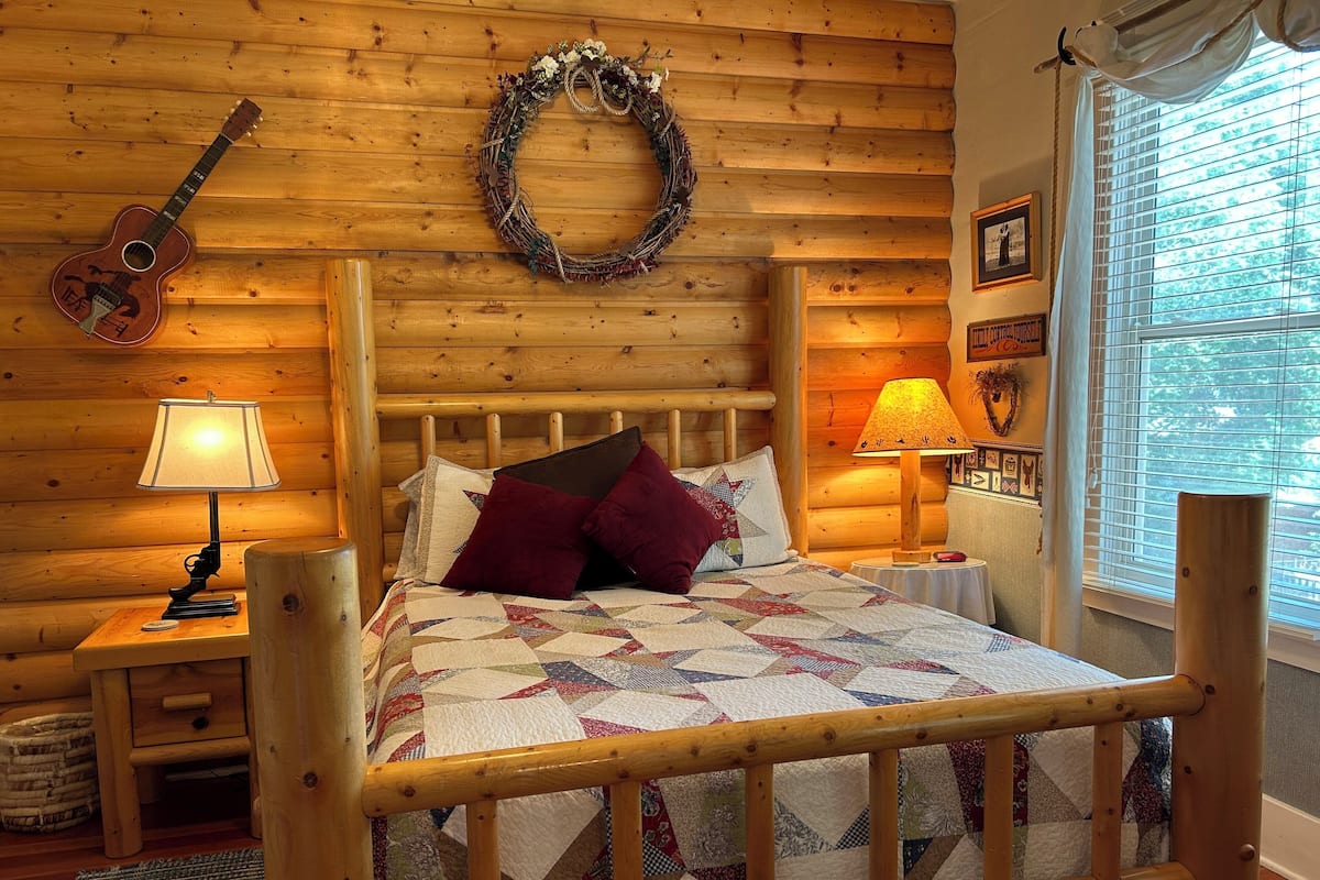 A cozy log cabin room with a wooden bed, two bedside lamps, a guitar, a wreath, and a window with blinds.