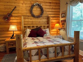 A cozy log cabin room with a wooden bed, two bedside lamps, a guitar, a wreath, and a window with blinds.