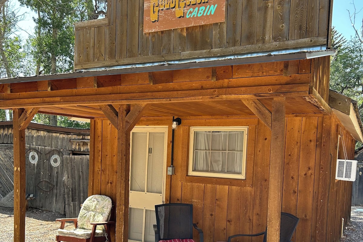 A wooden cabin with a sign reading "Goldilocks Cabin." It has a porch with two chairs and a small table.