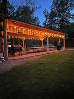 An outdoor covered picnic area with string lights, picnic tables, and a barbecue grill.
