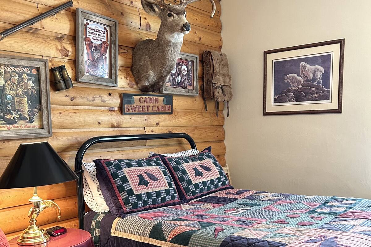 A cabin-themed bedroom with a bed, quilt, framed pictures, and a mounted deer head.