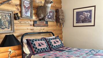 A cabin-themed bedroom with a bed, quilt, framed pictures, and a mounted deer head.