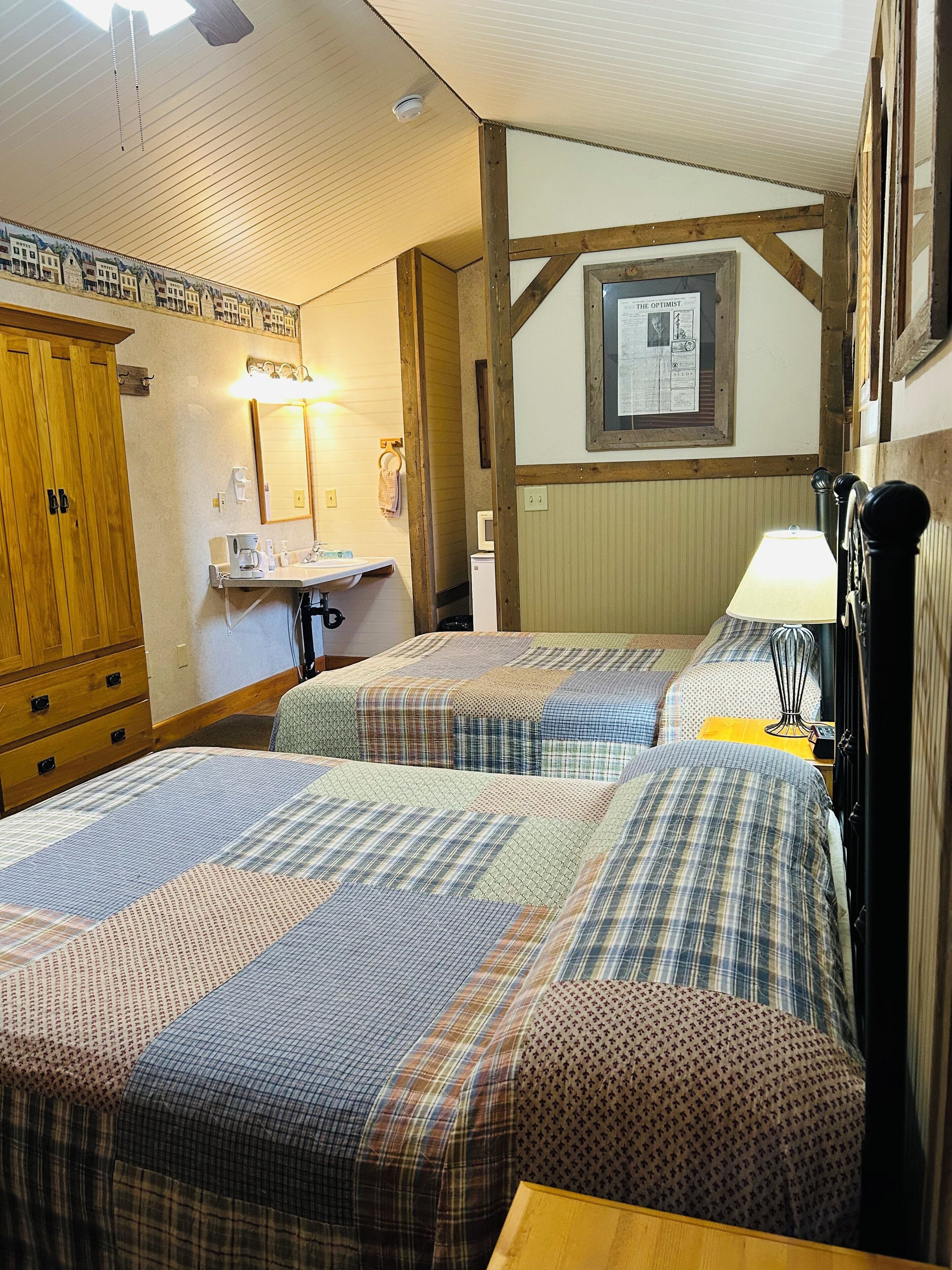 Ein Schlafzimmer mit zwei Betten, einem Holzschrank, einem kleinen Schreibtisch mit Lampe und einem an der Wand hängenden Bilderrahmen.