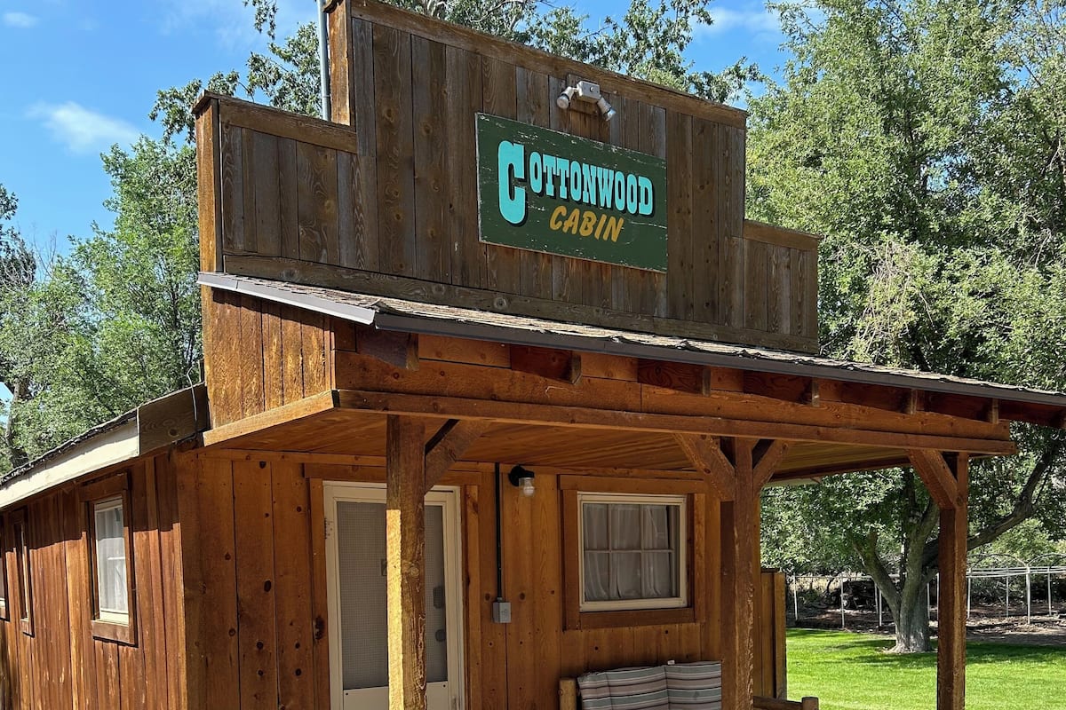A rustic wooden cabin labeled "Cottonwood Cabin" with a porch, a bench, and a small table with flowers.
