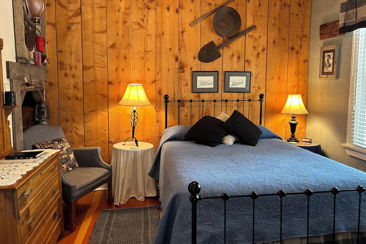 A cozy bedroom with a wooden wall, a bed with blue bedding, two bedside tables with lamps, a dresser, and a chair.