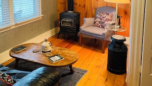 Un salon chaleureux avec un poêle à bois, un canapé confortable, une petite table avec un livre et une bouteille, et une lampe.