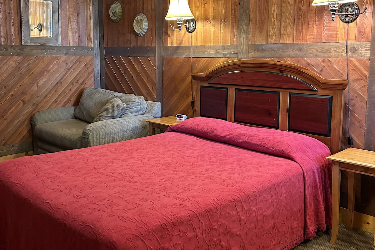 A room with a wooden wall, a bed with a red bedspread, a wooden nightstand, a chair, and a lamp.