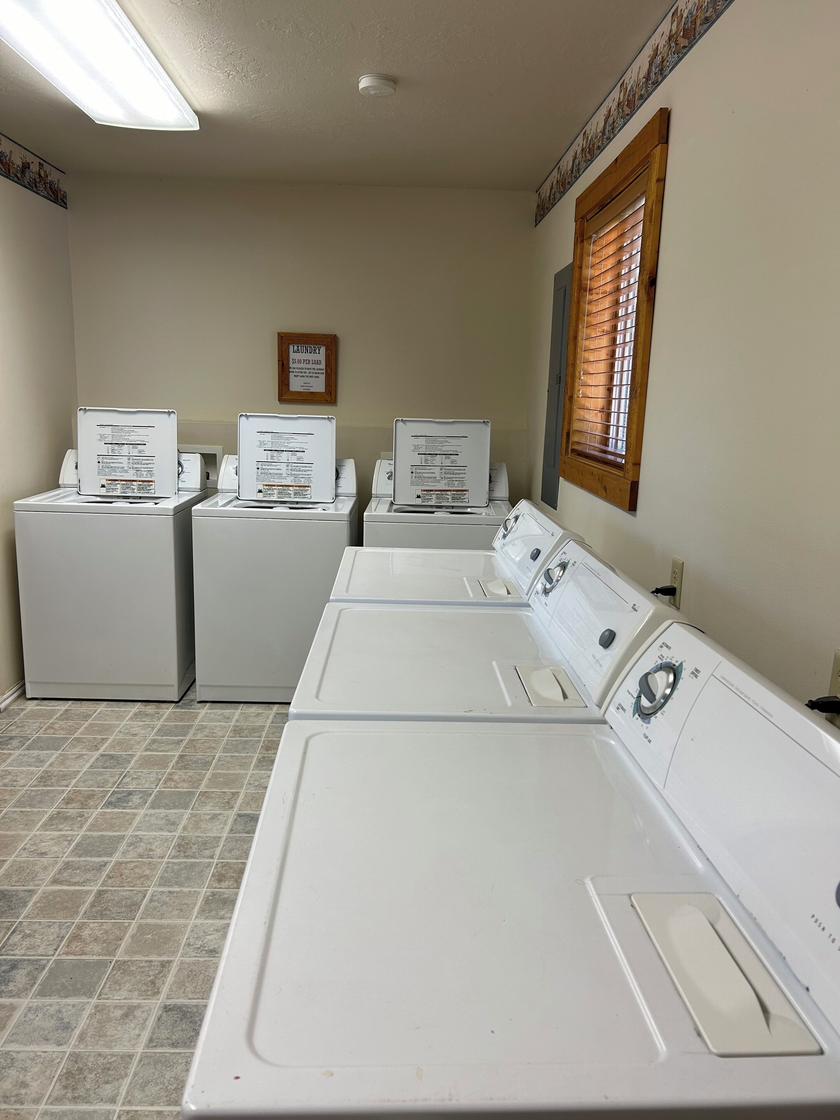 A laundry room with four front-loading washers and dryers, a ceiling light, a fire alarm, and a window with blinds.