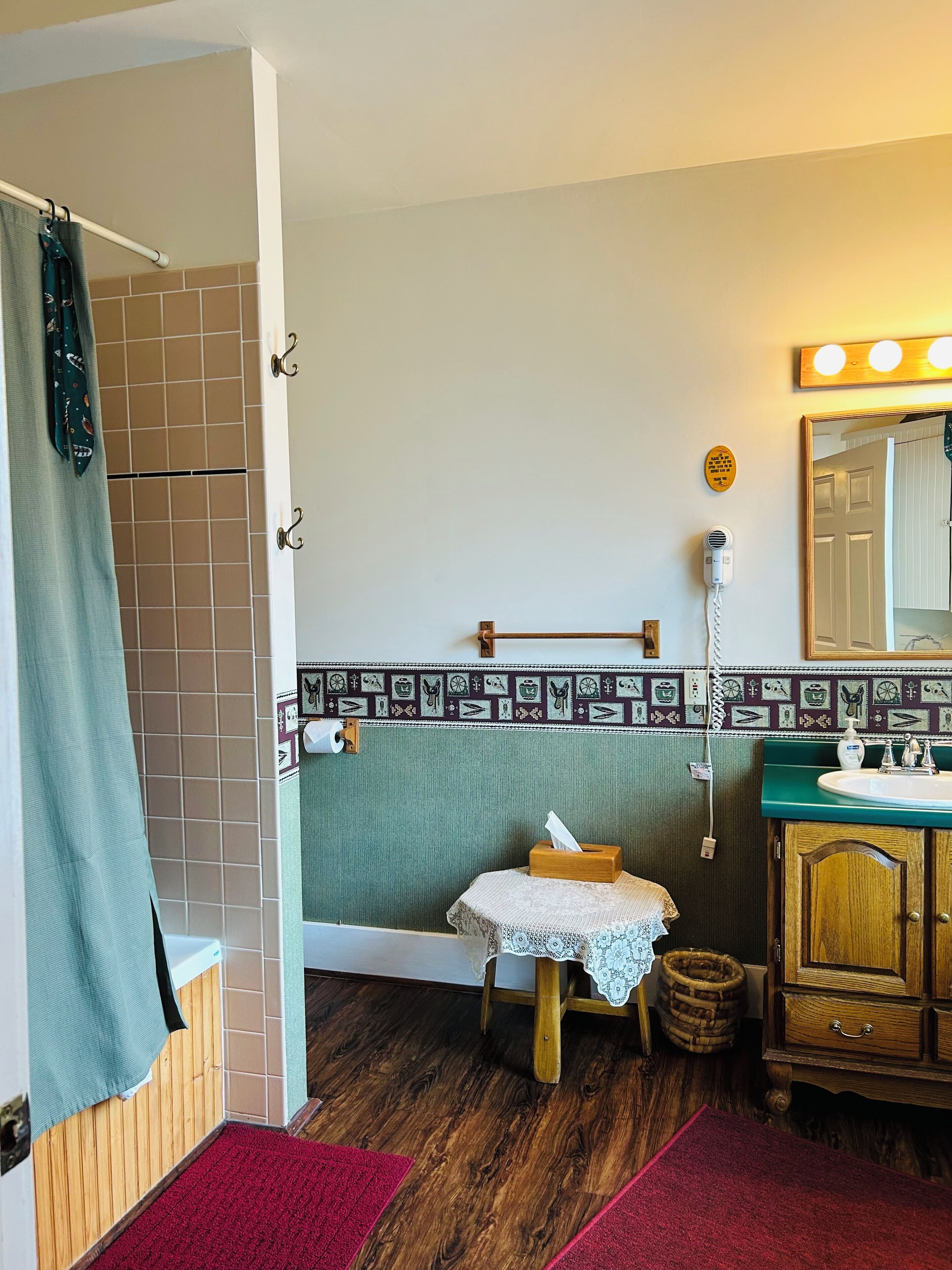 A bathroom with a wooden vanity, a mirror, a towel rack, a shower curtain, and a small table with a tissue box.