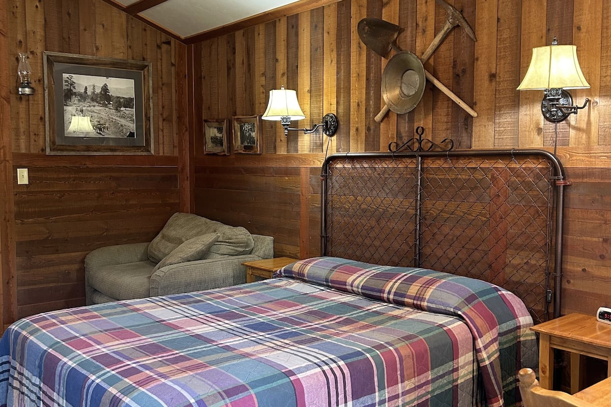A wooden cabin room with two beds, a couch, and a small table. There are wall-mounted lamps and a framed photograph.