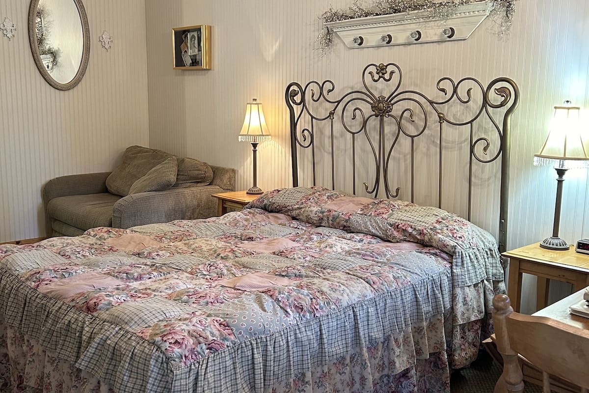 A bedroom with a floral bedspread, a wooden nightstand with a lamp, a cushioned chair, and a mirror on the wall.