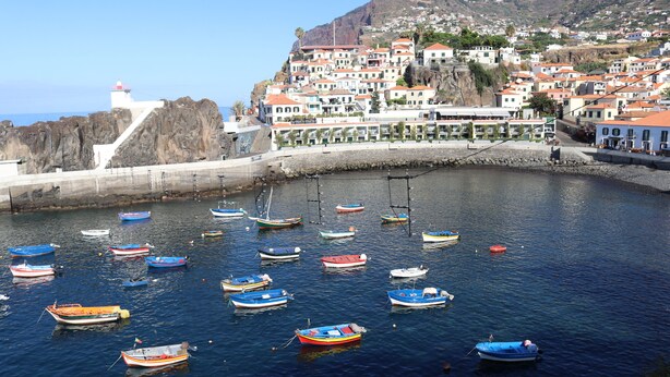Un poble costaner amb vaixells de colors amarrats en un port, un far i edificis amb teulades vermelles.