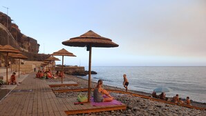 Una terrassa de fusta amb sombrerets de palla, una platja rocosa i mar tranquil·la.