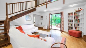 A spacious living room with a wooden staircase, a white sofa, a red ottoman, a bookshelf, and a view of greenery through a glass door. - Gorgeous home in Malinska (Malinska)