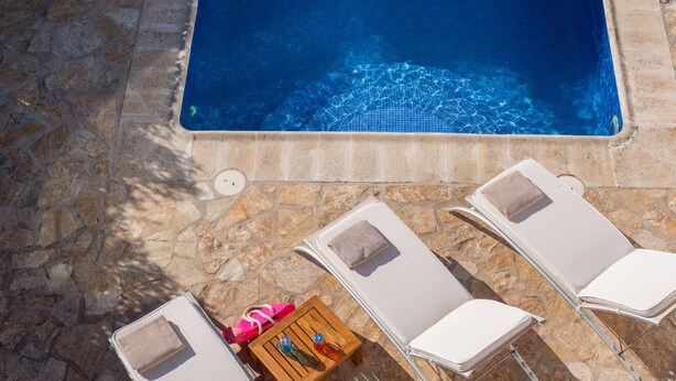 A pool area with three sun loungers, a small wooden table with a bottle and a pink bag, and a stone tiled floor. - Awesome home in Ibiza (Ibiza)
