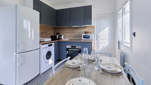 A compact kitchen with a washing machine, microwave, and oven. A dining table with plates and glasses is set for six. - Appartement Évasion Normande - Welkeys (Cabourg)
