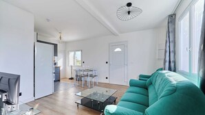 A modern living room with a teal sofa, a glass coffee table, and a kitchen area in the background. - Appartement Évasion Normande - Welkeys (Cabourg)