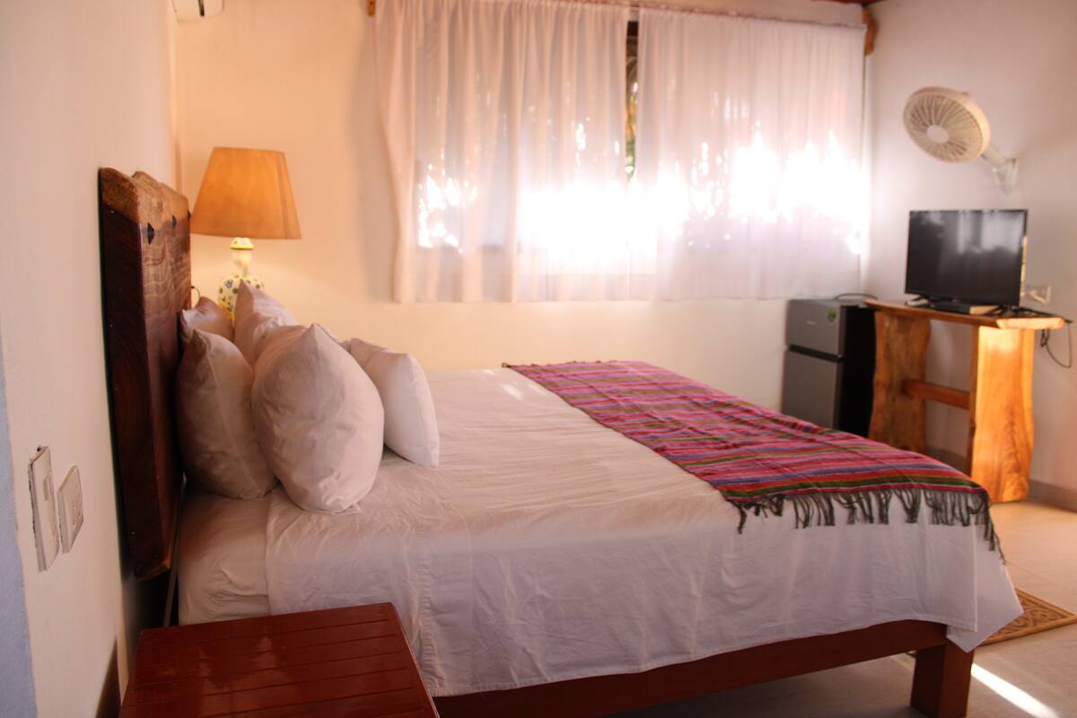 A bedroom with a bed, a desk with a computer, a lamp, and a fan.