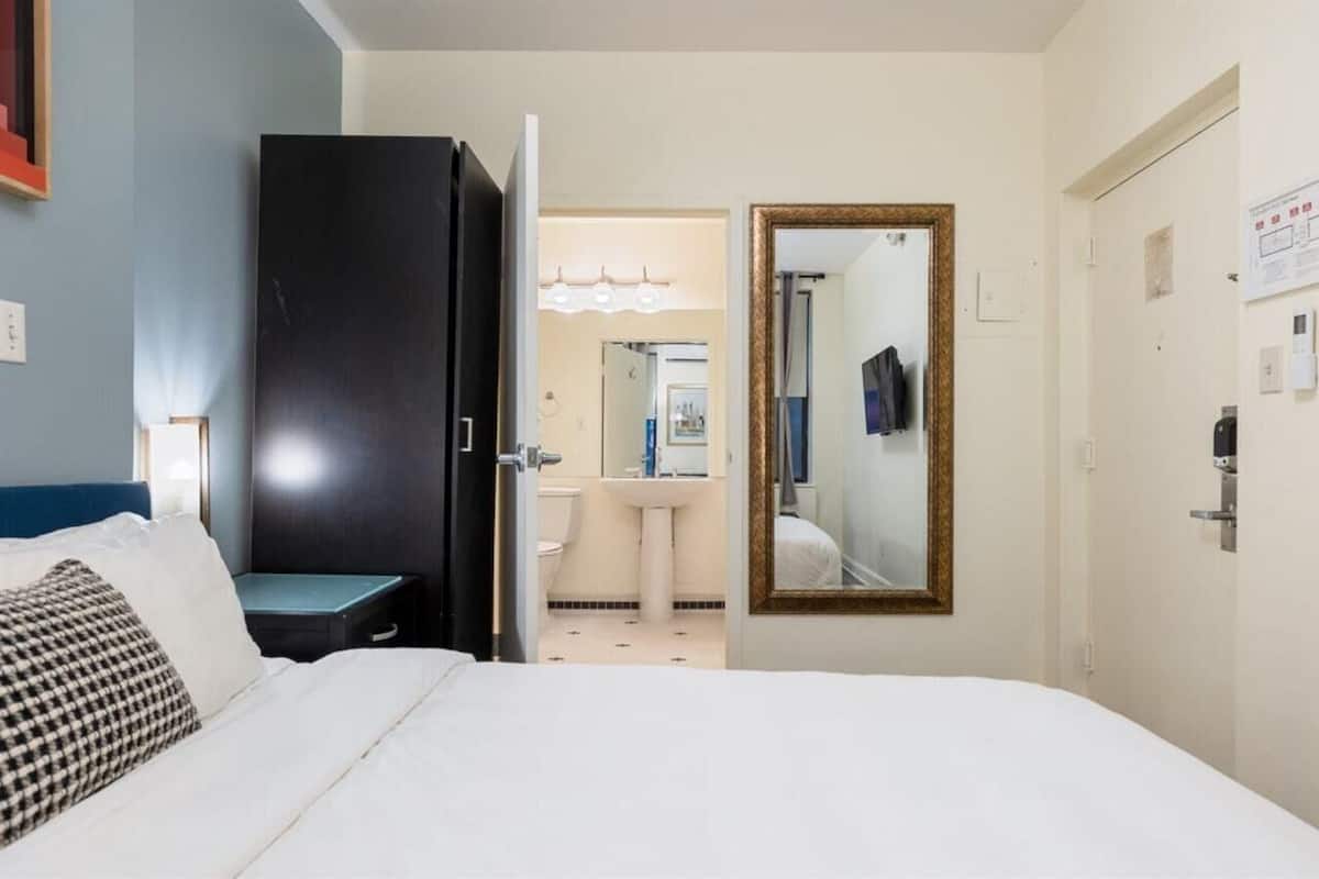 Habitación de hotel con cama, armario, baño con lavamanos y espejo, y una puerta que da a otra habitación.