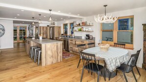 A spacious kitchen with wooden cabinets, a central island, and bar stools. There is a dining area with a table and chairs, and a chandelier above. - Lincoln Lodge! Steps from the heart of Elk Avenue! (Crested Butte)