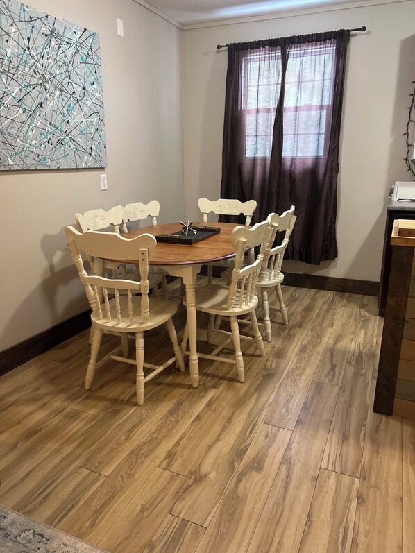 A dining area with a wooden table and chairs, a painting on the wall, and a window with curtains. - The Rustic Oasis (Kentville)