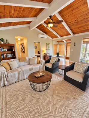 A spacious living room with a high ceiling, wooden beams, a large sofa set, a coffee table, and a bookshelf.