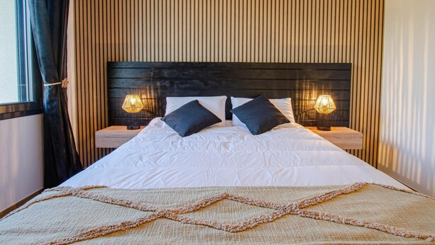 Un lit bien fait, avec des draps blancs et deux oreillers foncés, flanqué de deux tables de chevet avec des lampes, le tout adossé à une tête de lit en bois et placé devant un mur à rayures.