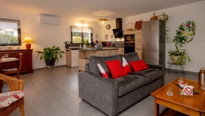 A modern living room with a grey sofa, a dining area with a table and chairs, a kitchen with a fridge, and a window with a view. - Holiday Home 'Les Chaudannes' with Mountain View, Wi-Fi, and Air Conditioning (Valgelon-La Rochette)