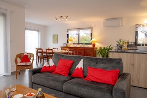 A modern living room with a grey sofa, red cushions, a dining area with wooden chairs, and a kitchen counter with a plant. - Holiday Home 'Les Chaudannes' with Mountain View, Wi-Fi, and Air Conditioning (Valgelon-La Rochette)