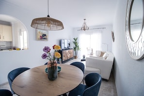 Uma área de refeições com mesa redonda de madeira, cadeiras azuis, um vaso com flores e uma luminária trançada pendente.