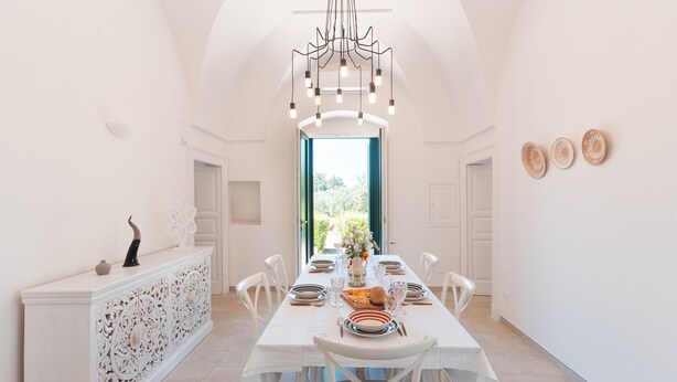 Un espace repas avec une longue table, des chaises blanches, un lustre et une vue sur l’extérieur par une porte ouverte.
