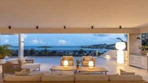 Un área de descanso moderna al aire libre con vista al mar, que incluye sofás, mesas e iluminación ambiental.
