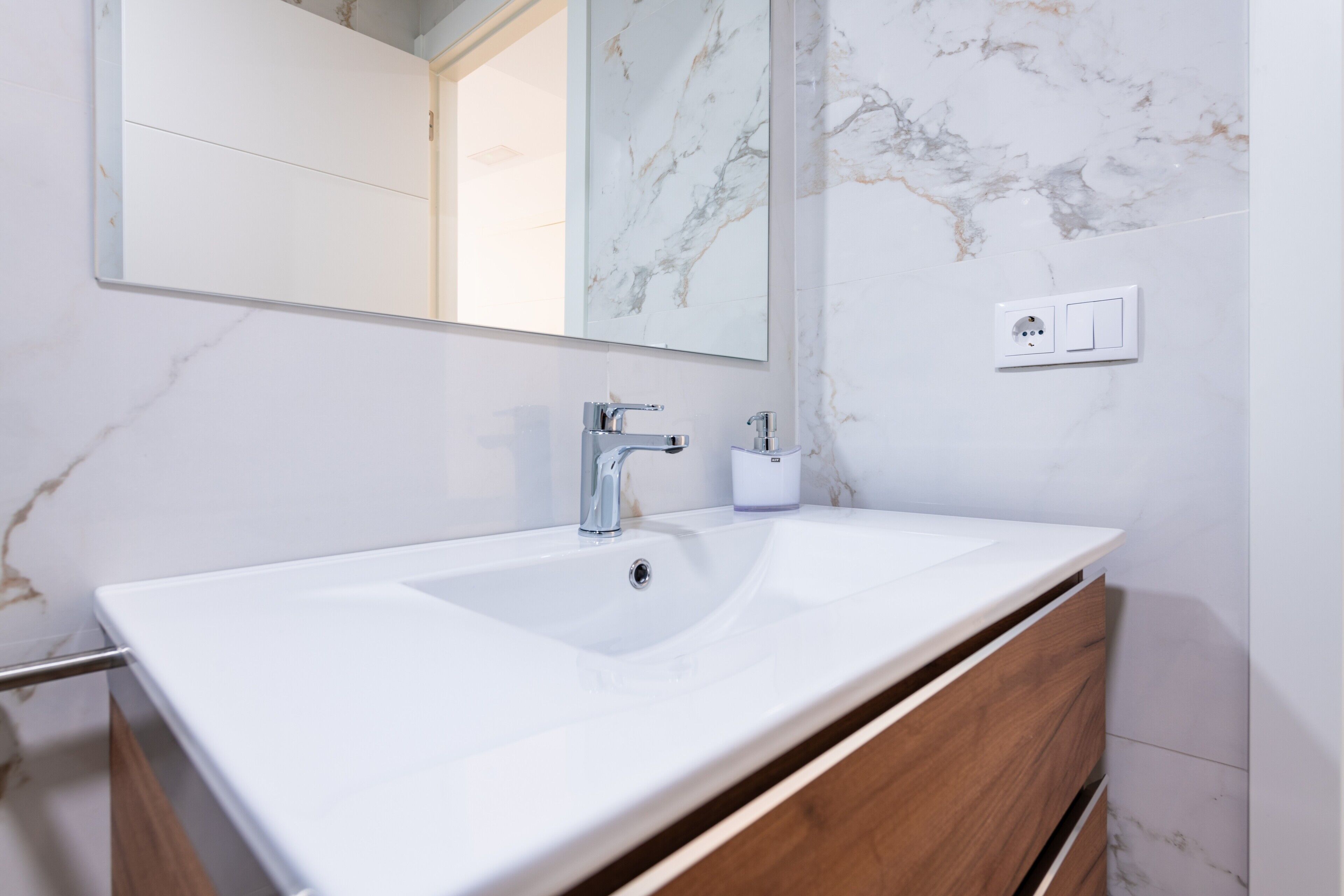 Un baño con lavamanos blanco, espejo y pared de mármol.