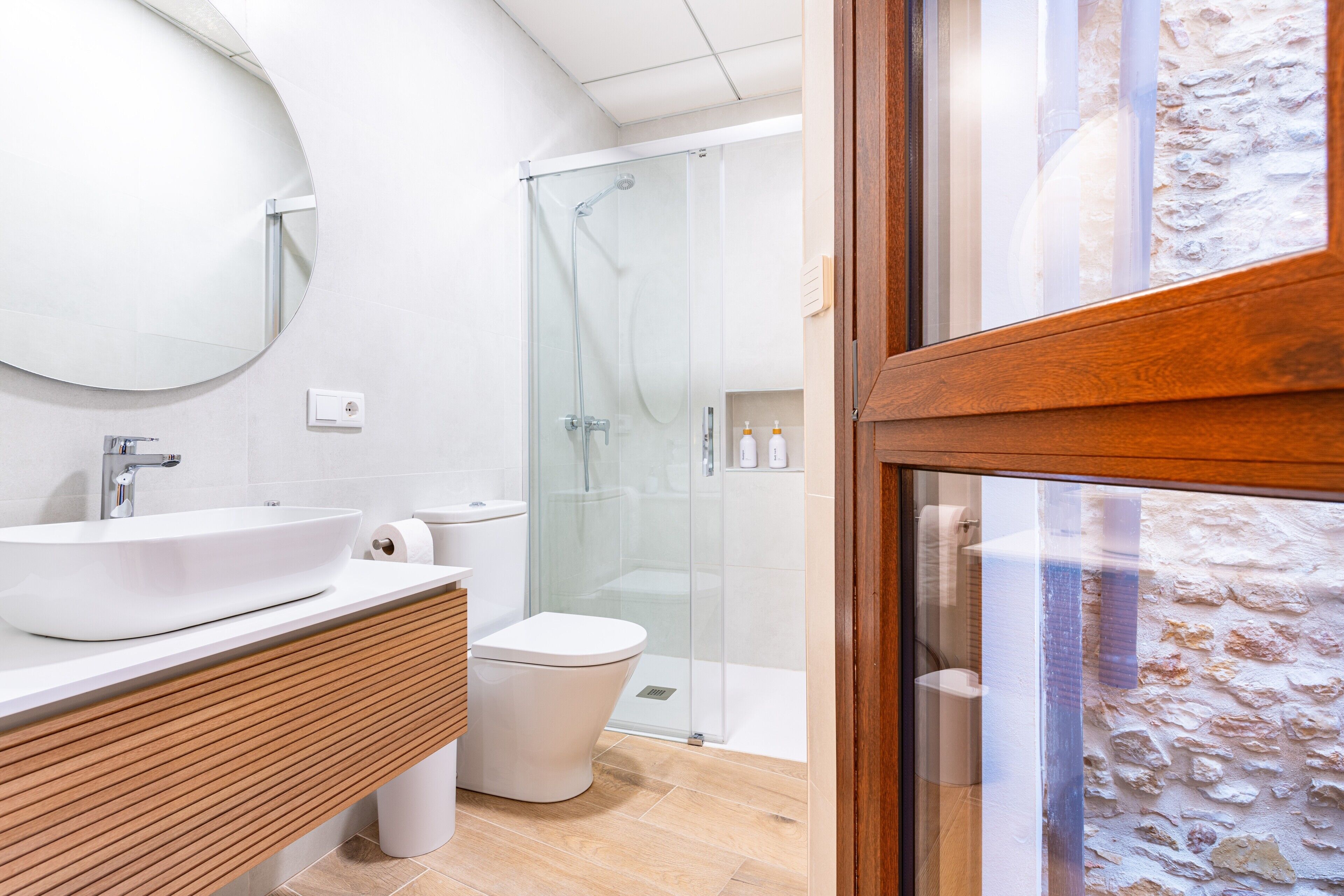 Et moderne badeværelse med et rundt spejl, hvid vask, trækommode, toilet og en brusekabine med glasvægge.