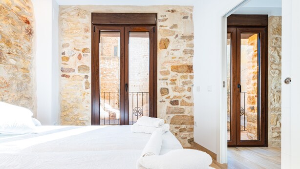 Un dormitorio con pared de piedra, una cama con ropa de cama blanca y dos puertas de madera que dan a un balcón.