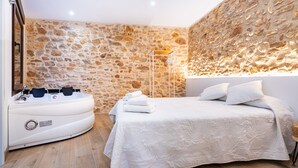 Un dormitorio con pared de piedra, una cama con ropa de cama blanca, una bañera blanca con controles y una lámpara de pie.