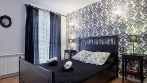 A bedroom with a bed, a nightstand, a lamp, a wall clock, and a window with curtains.