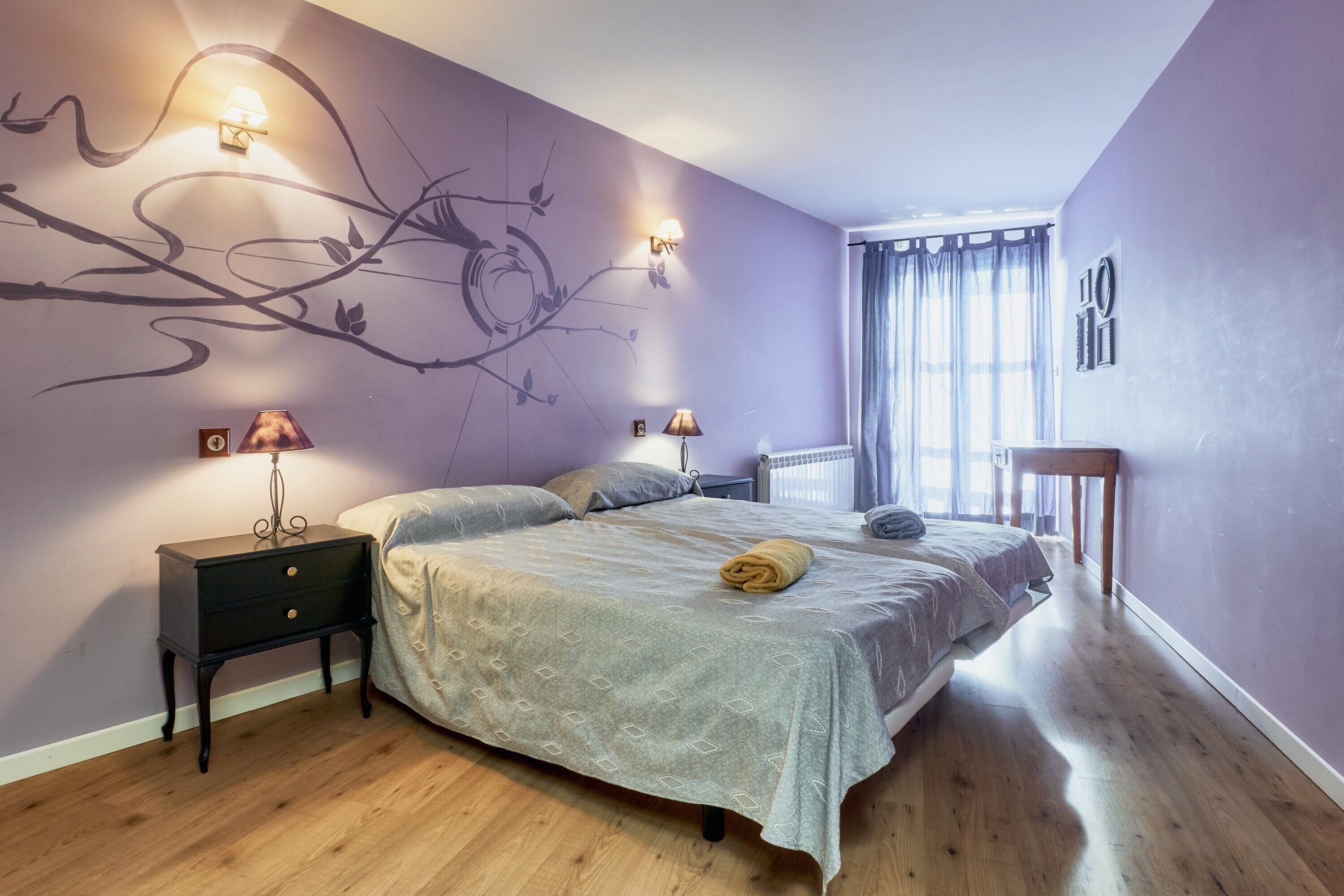 Un dormitorio con cama, dos mesitas de noche con lámparas, un mural en la pared y una ventana con cortinas.