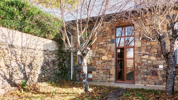 A stone building with a large window and a door, surrounded by trees and a stone wall.