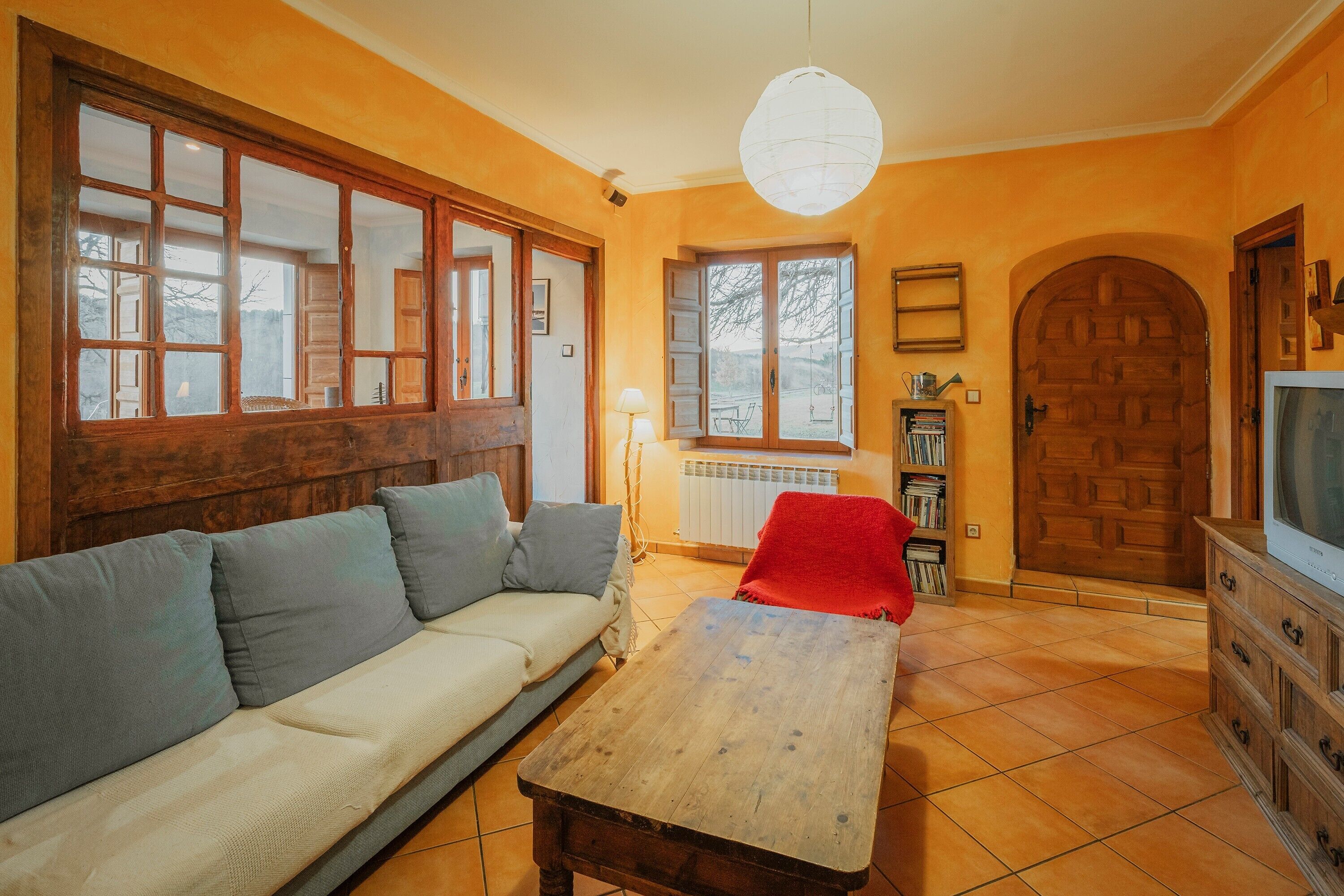 Un salon comprenant un canapé, une table basse en bois, un plaid rouge, un téléviseur, une bibliothèque et une grande porte en bois.