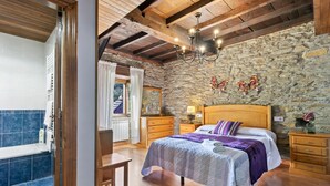 Quarto com parede de pedra, vigas de madeira no teto, cama com colcha roxa, mesa de cabeceira com abajur e uma cadeira de madeira.