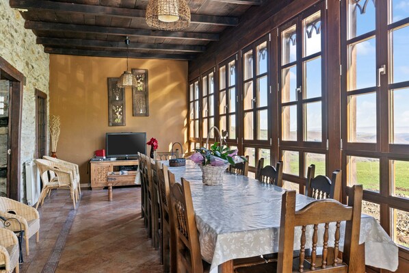 A dining area with a long table, chairs, a television, and a view through large windows.