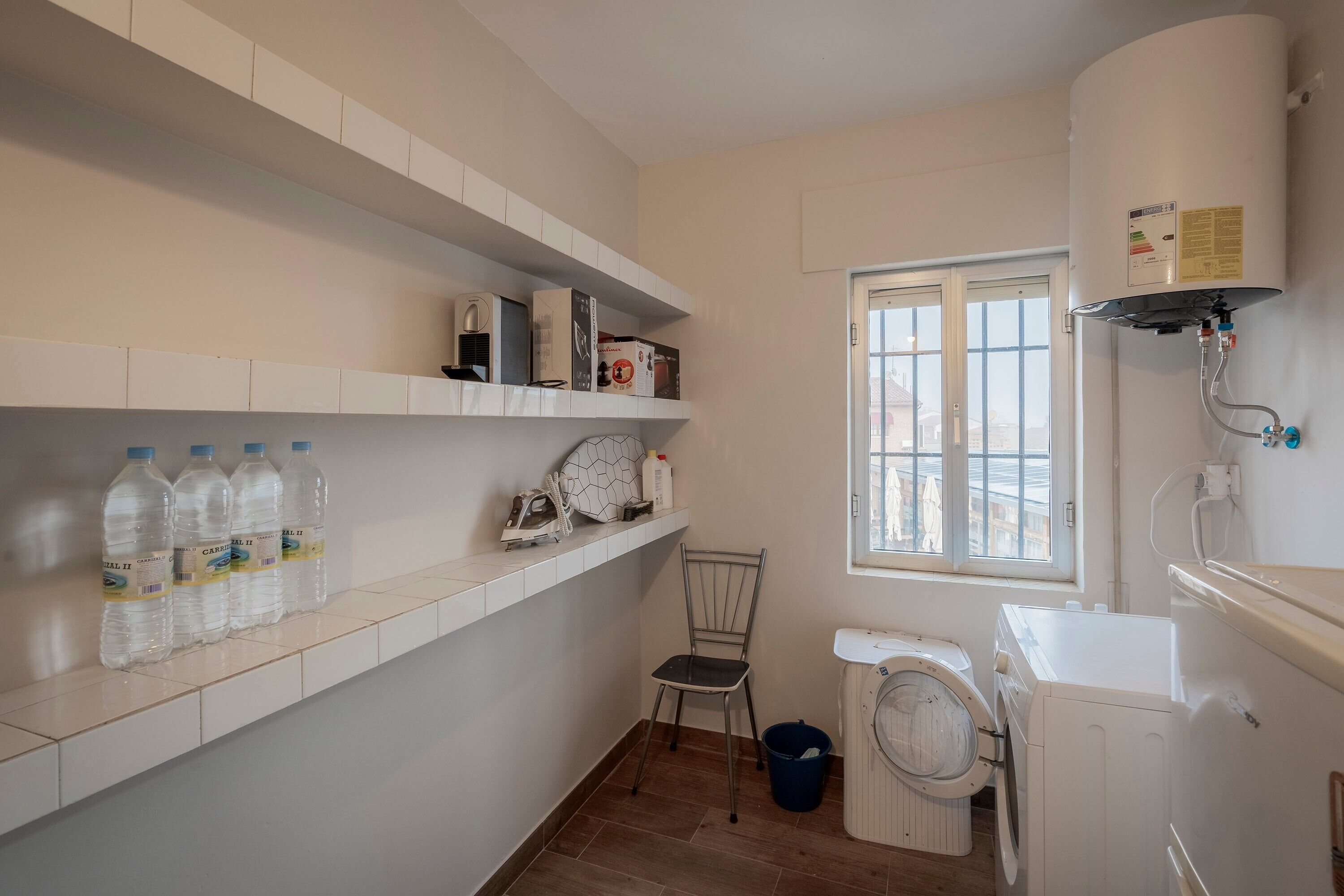 A laundry room with a washing machine, dryer, and a water heater. There are shelves with bottles and a chair.