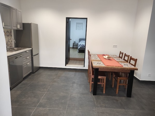 A compact kitchen with modern appliances, a dining area with a wooden table and chairs, and a view into a bedroom through an open door.